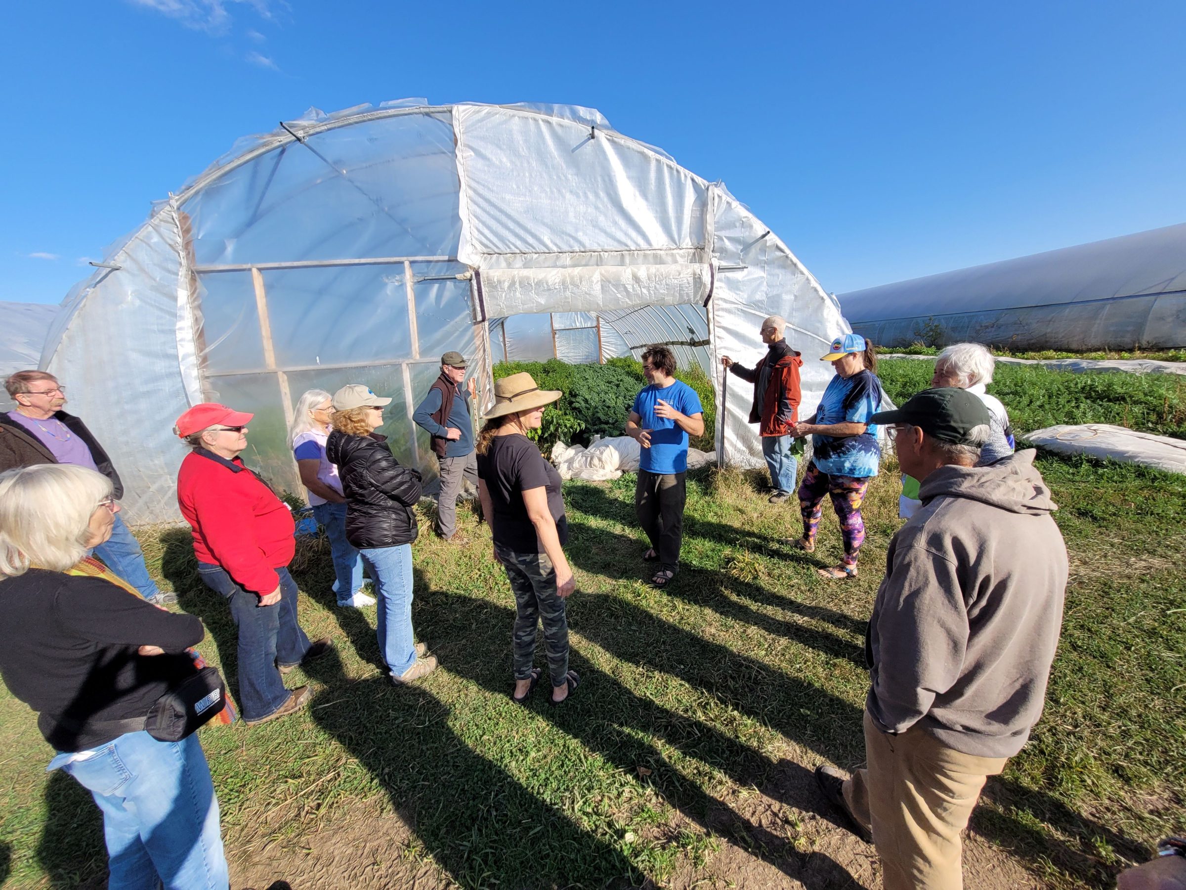 A group of people stand in front of a greenhouse with a man in the middle of the group talking.