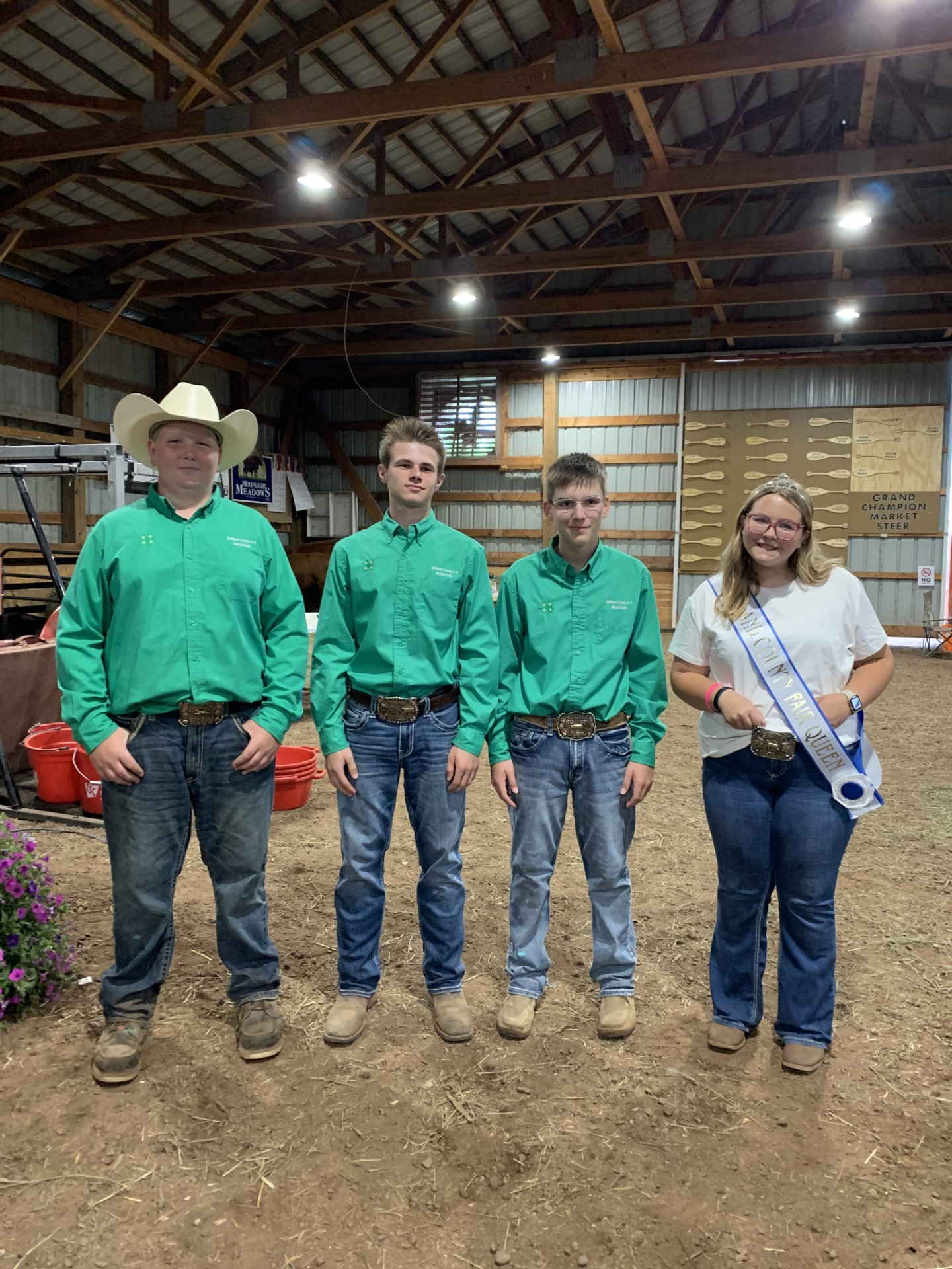 A group of four youth standing in a barn showing off their belt buckles for 4-H. Three are in matching green shirts and one is in a white shirt with a crown and sash.