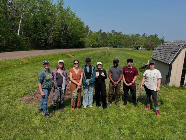 A group of 8 people standing in front of a garden bed in the middle of a field of short grass. It is a sunny day and most people are in hats or sunglasses.