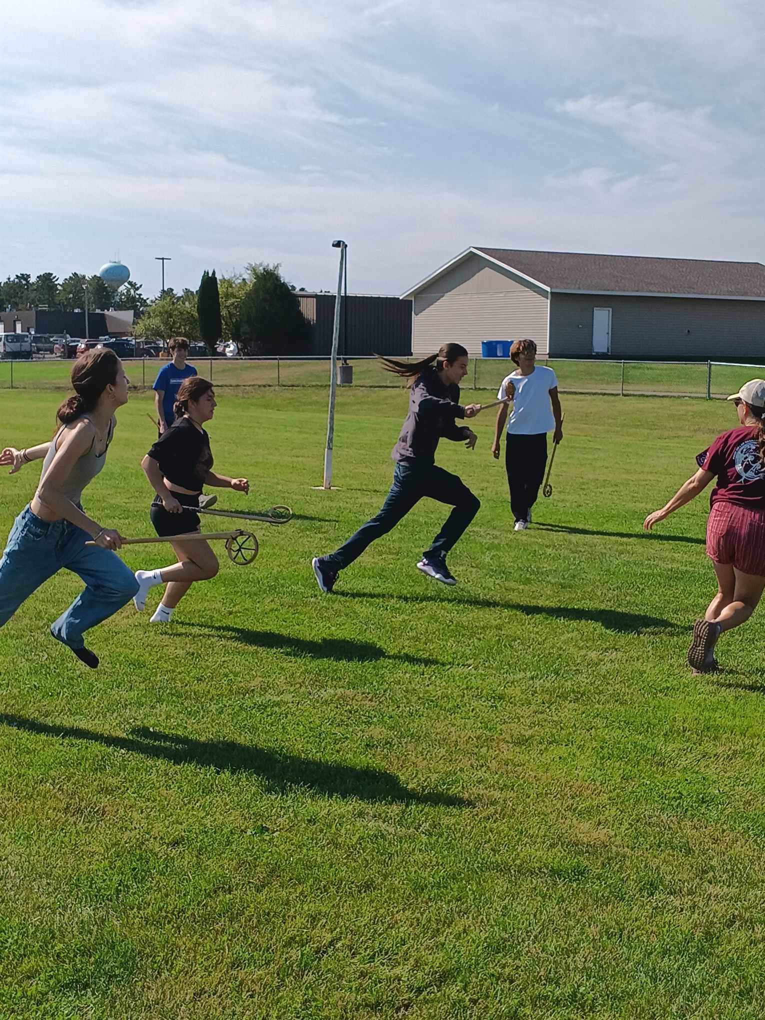 Four people running in a grassy field holding lacrosse sticks. They are running from the left side of the frame to the right side.