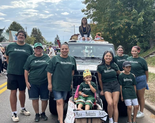 A group of youth and adults in matching green t shirts stand smiling at the camera waiting for a parade to start.