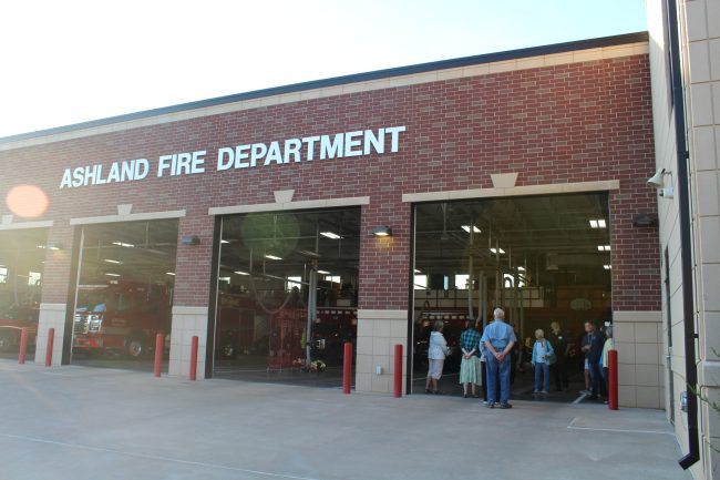 a brick building with the words Ashland Fire Department and large garage doors that are open. A group of people stand just inside one of the garage doors.